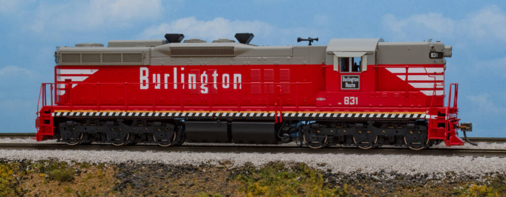 Color photo of HO scale diesel locomotive painted red and gray with white, yellow, and black graphics on scenicked base with a sky blue backdrop.
