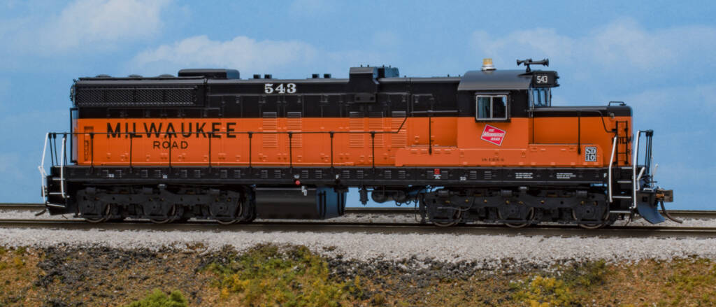 Color photo of HO scale chopped-nose locomotive with single fuel tank painted orange and black on scenicked base with a sky blue backdrop.