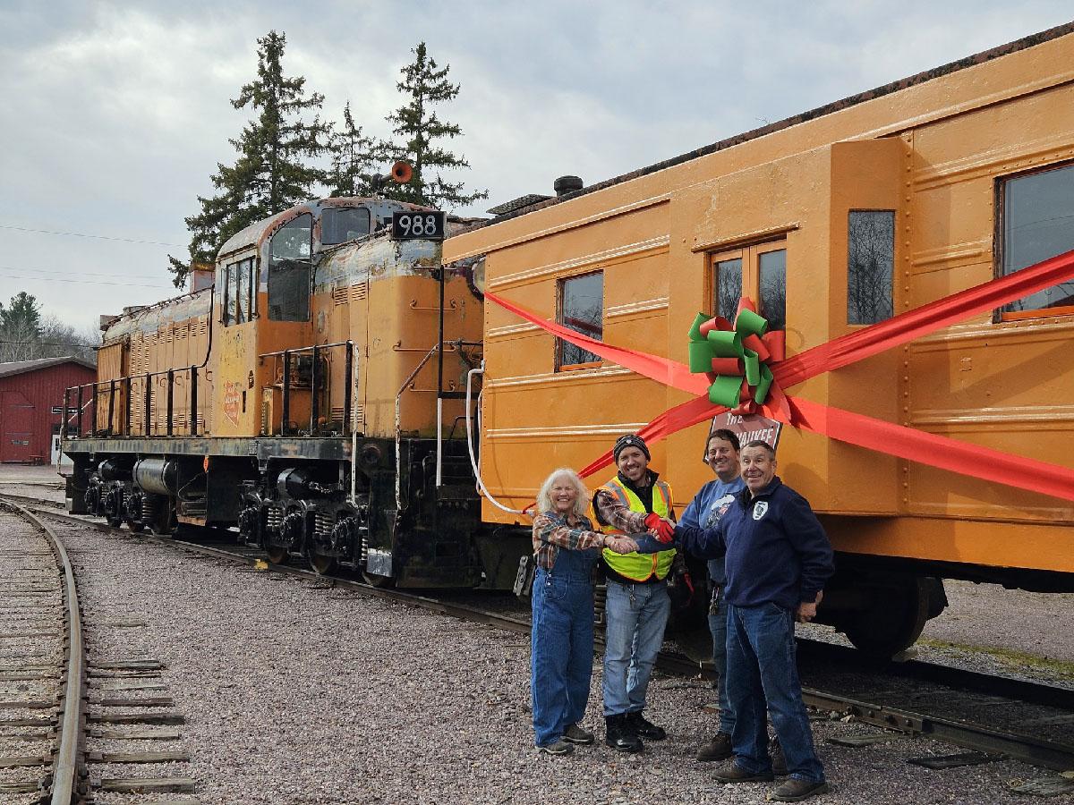 Orange and gray diesel locomotive with an orange caboose and four people shaking hands. Milwaukee Road caboose donated.