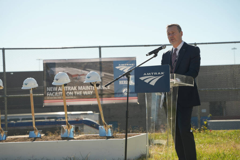 Man speaking at podium outdoors with shovels in background