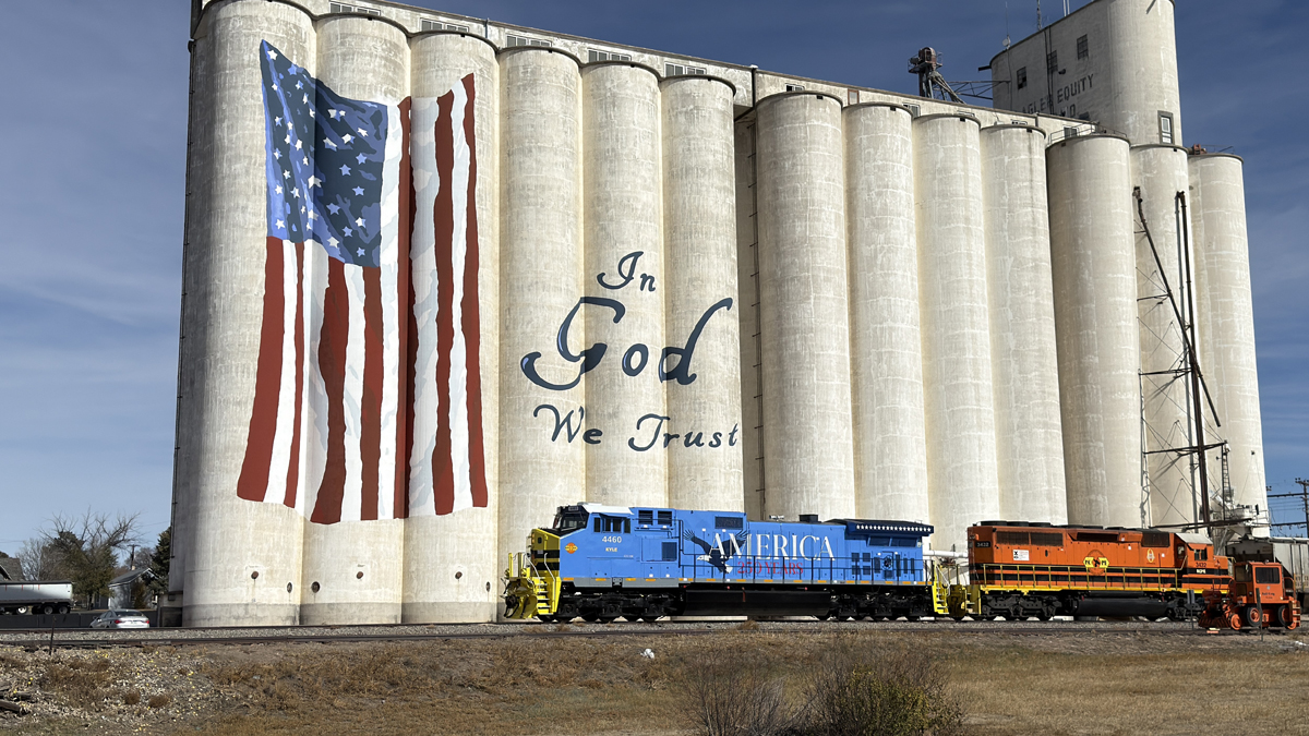 Light blue "America 250" locomotive passes grain elevator painted with American flag and "in God We Trust" logo