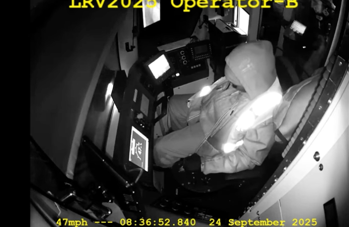 Black-and-white image from in-cab camera showing operator of light rail vehicle with head down, chin on chest
