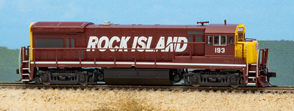 Color photo of four-axle N scale diesel locomotive painted maroon and yellow with white graphics on scenicked base with a sky backdrop.