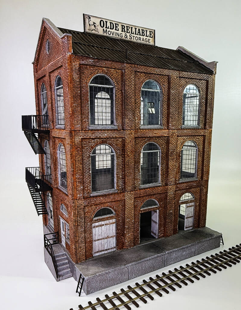 Color photo of HO scale two-story brick warehouse with peaked roof, black-and-white signs, large windows, and track dock with section of track on white background.