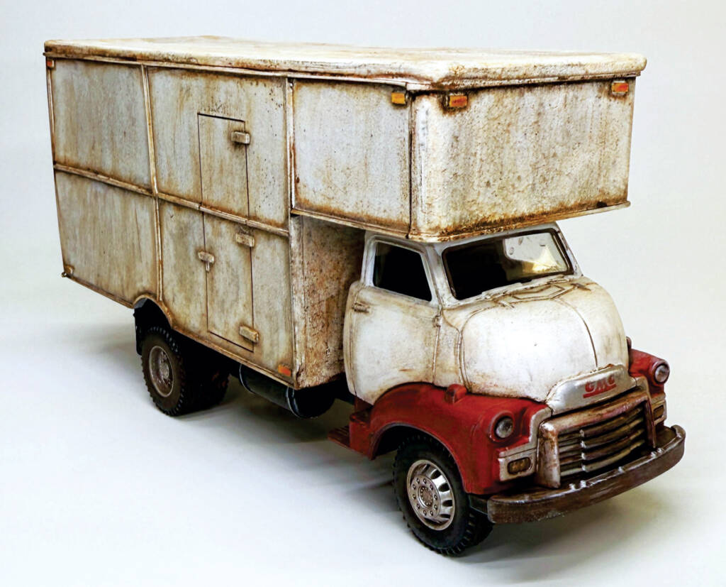 Color photo of O scale moving truck with weathered white body, weathered red-and-white cab, and weathered tires on white background.