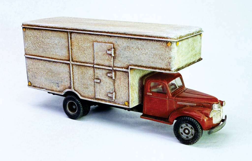 Color photo of HO scale moving truck with weathered white body, weathered red cab, and weathered tires on white background. 