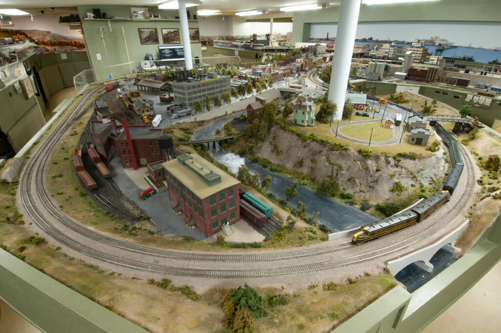 Color photo of HO scale model railroad with structures, roads, tracks, and rolling stock; two support columns; and wall in background with three photos.