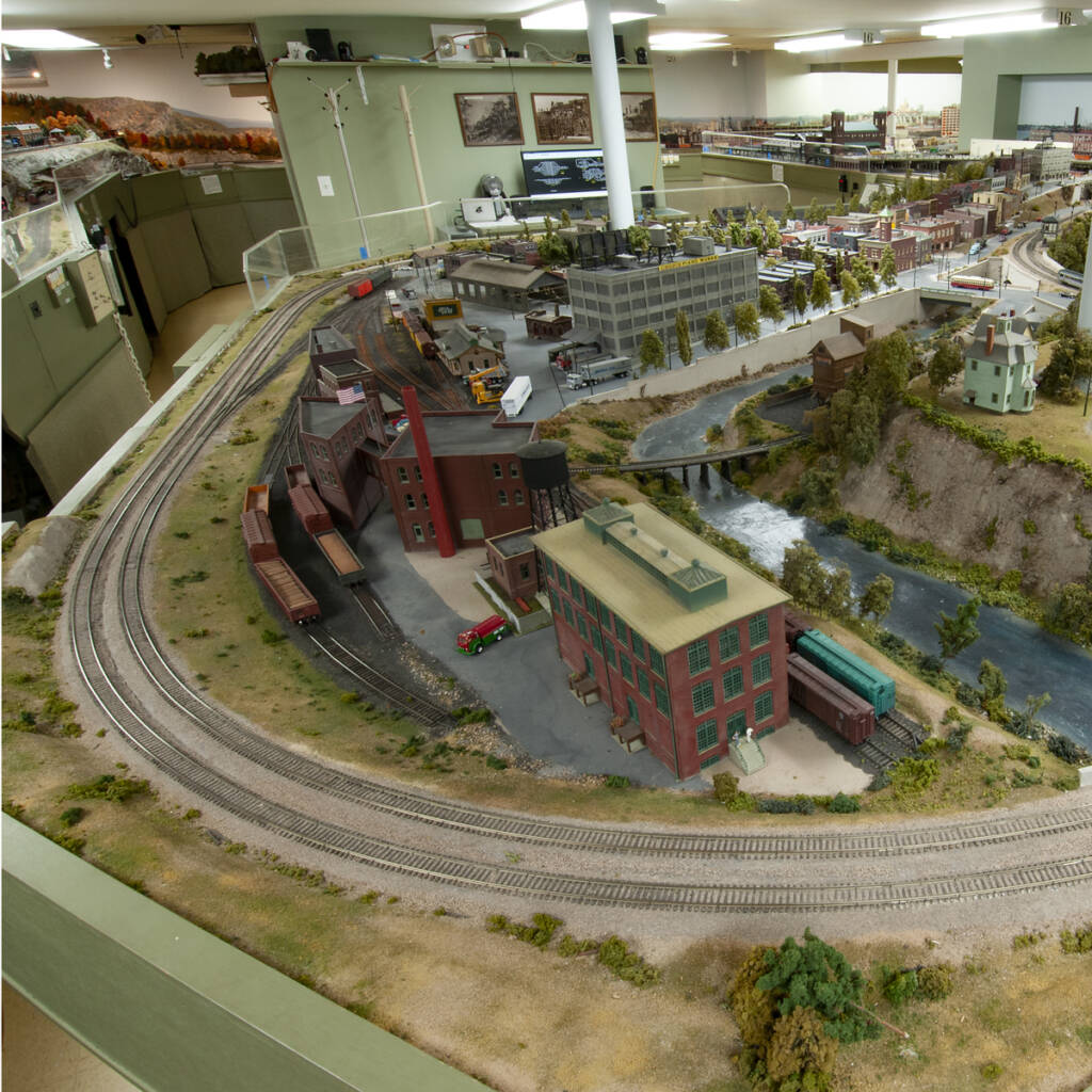 Color photo of HO scale model railroad with structures, roads, tracks, and rolling stock; two support columns; and wall in background with three photos.