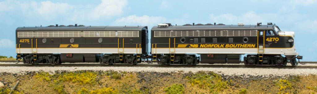 Color photo of HO scale four-axle EMD cab and cabless units painted black, white, and gold with gold graphics on scenicked base with a sky blue background. 