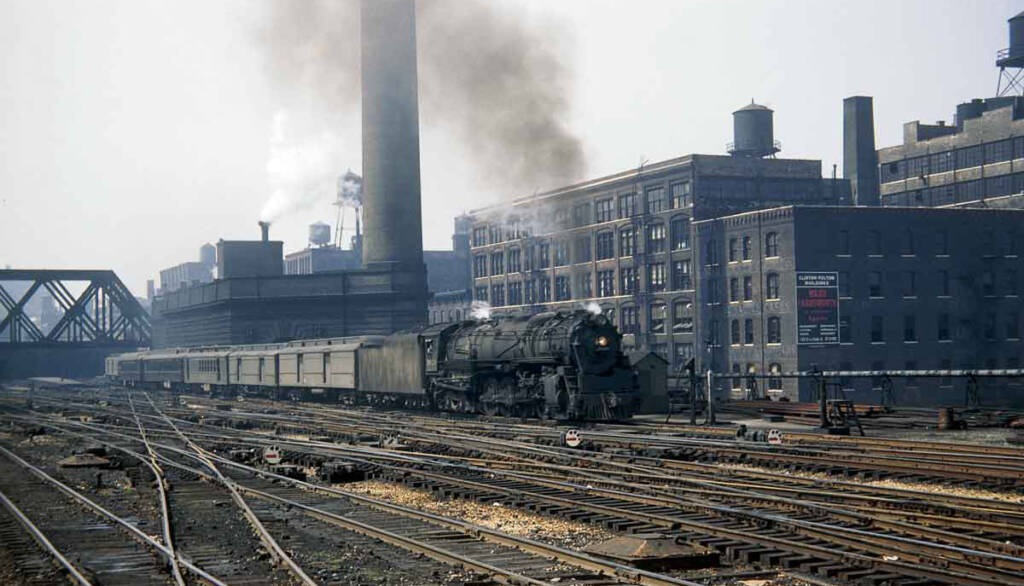 A vintage color photograph of a steam locomotive in an urban train yard