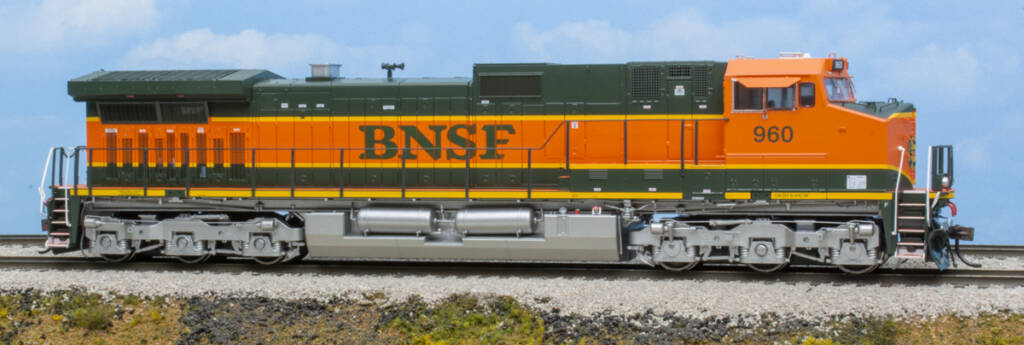 Color photo of modern HO diesel painted green, orange, and yellow with silver trucks and fuel tank on scenicked base with a sky and clouds in background.