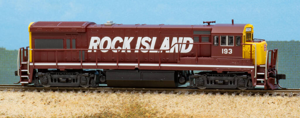 Color photo of N scale four-axle diesel locomotive painted maroon, yellow, and black with white graphics on scenicked base with a photo backdrop.