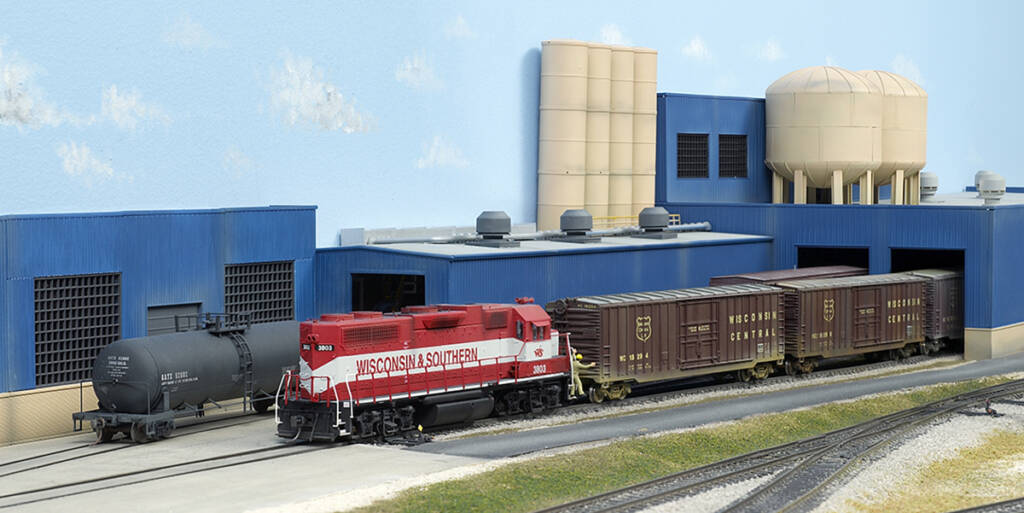 Color photo of red and gray HO scale EMD hood unit switching maroon boxcars at tan and blue industry with faded black tank car in background.
