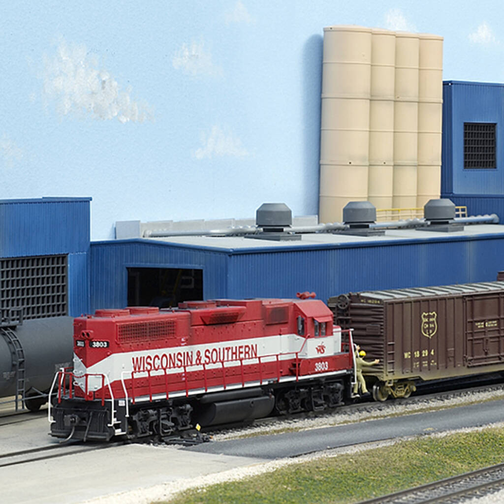 Color photo of red and gray HO scale EMD hood unit switching maroon boxcars at tan and blue industry with faded black tank car in background.