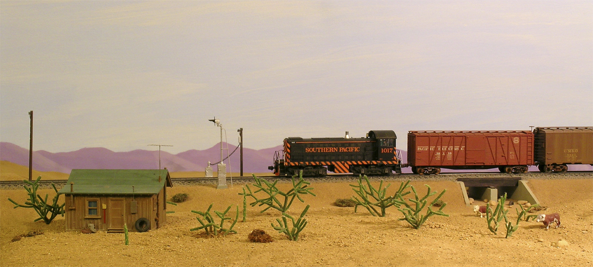 A black and orange model locomotive leads a train with orange boxcars through a desert model railroad layout, with live cactus plants in the foreground