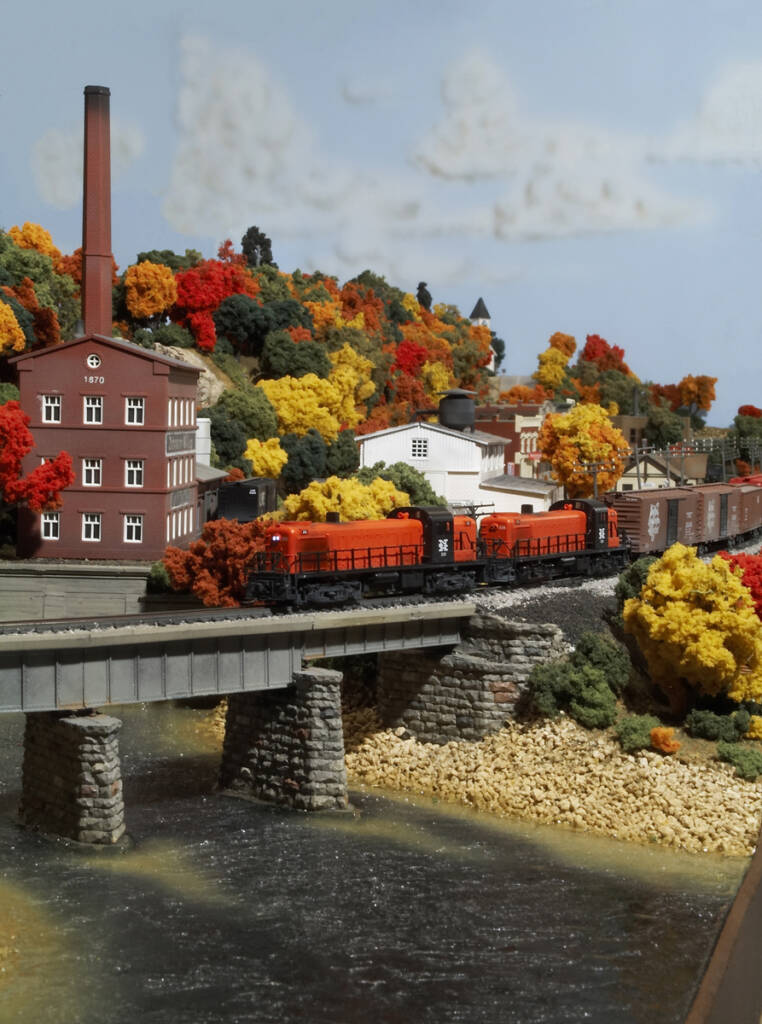 An orange model locomotive leads a train over a bridge on a model railroad layout with an autumnal setting
