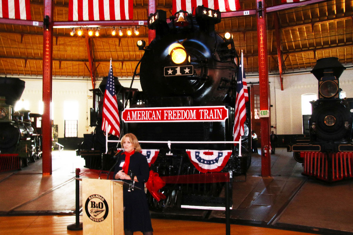 Lady standing at podium in front of black steam locomotive in a roundhouse.