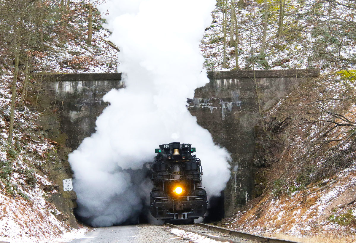 Steam locomotive emerges from tunnel on a cold day, clouded by steam.