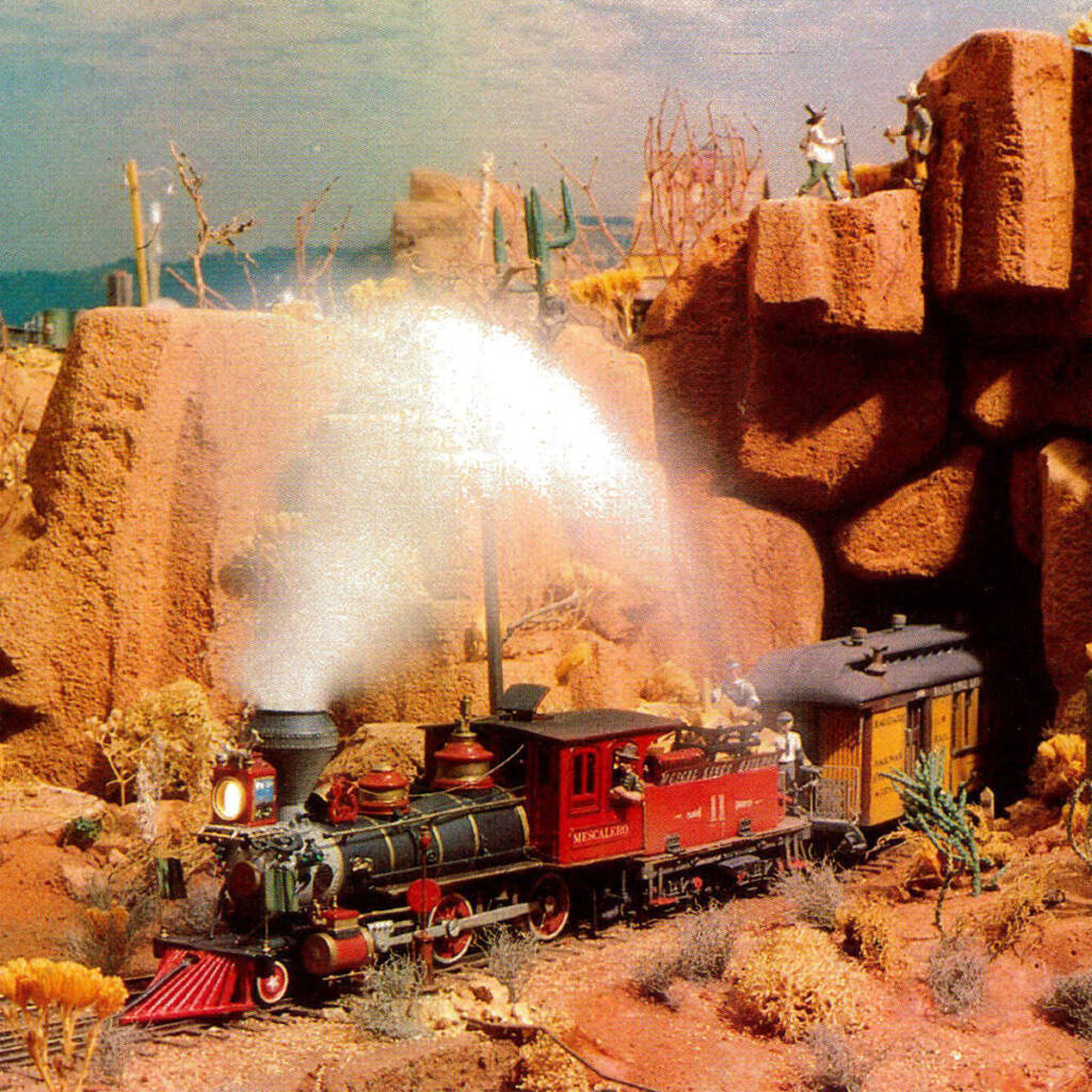 Color photo of red and black steam locomotive pulling yellow passenger car with black roof through Desert Southwest scenery.