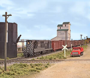A black locomotive pulls a string of boxcars toward a track diamond with a tan wooden grain silo in the background