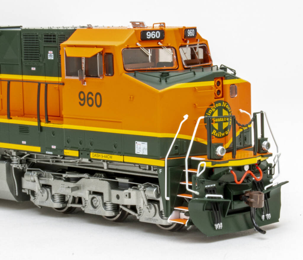 Color photo showing cab on an HO scale modern GE six-axle diesel locomotive painted orange, green, and yellow with silver trucks and fuel tank on white background.