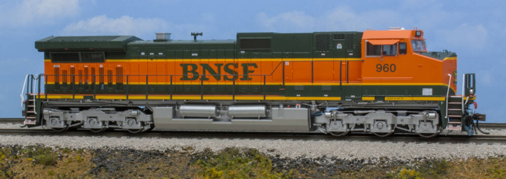 Color photo of HO scale modern GE six-axle diesel locomotive painted orange, green, and yellow with silver trucks and fuel tank on scenicked base with a sky blue background.