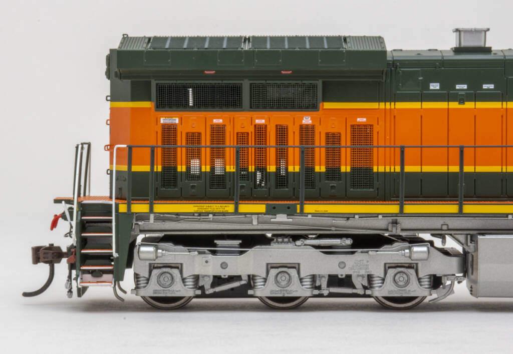 Color photo showing radiator section on an HO scale modern GE six-axle diesel locomotive painted orange, green, and yellow with silver trucks and fuel tank on white background.