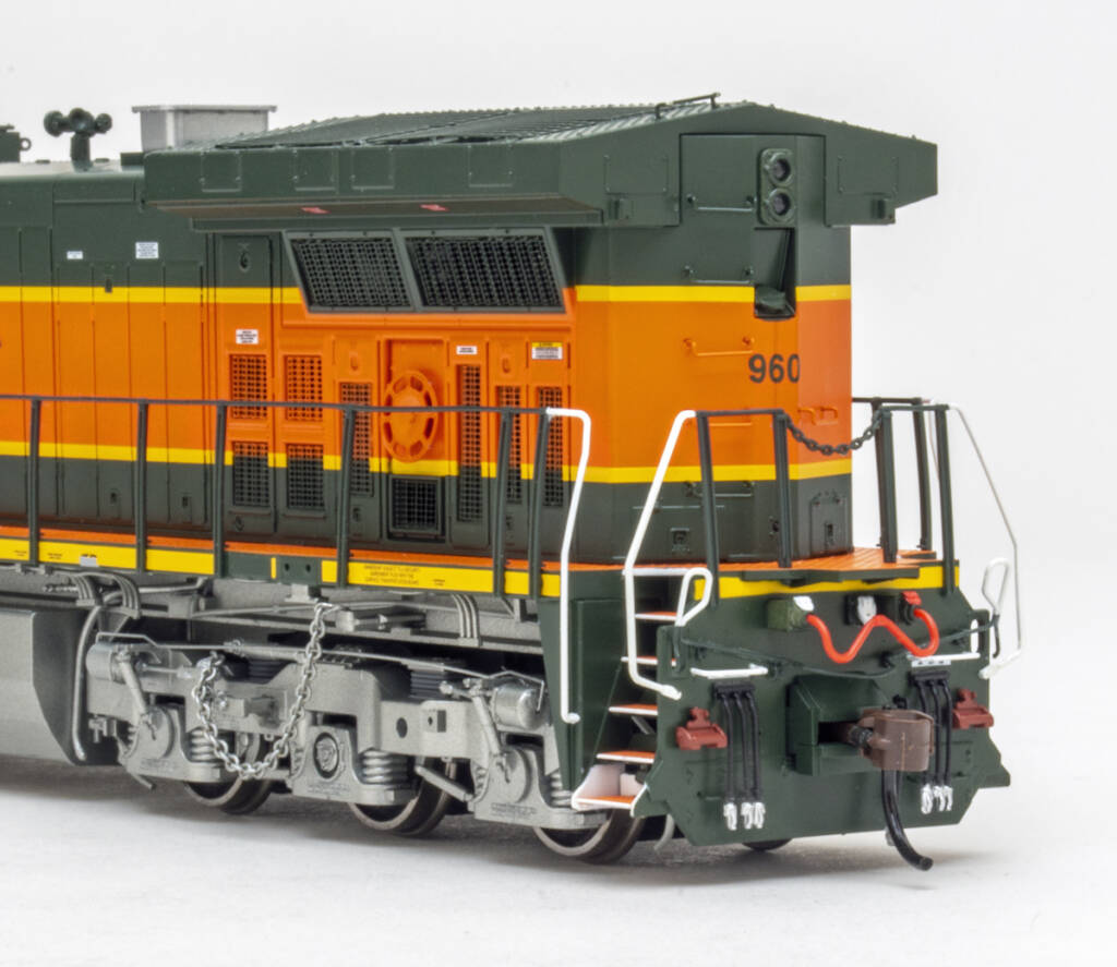 Color photo showing fireman’s side and end of long hood on an HO scale modern GE six-axle diesel locomotive painted orange, green, and yellow with silver trucks and fuel tank on white background.