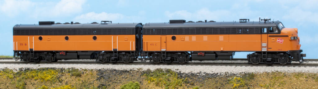 Color photo of two HO scale hood units painted orange and black with red and white graphics on scenicked base with a sky blue backdrop. 