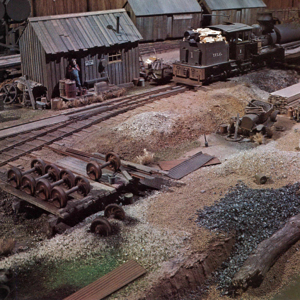 Color photo of scene on a model railroad that includes geared steam locomotive, weathered wood sheds, painted figure, wheelsets, and rusty debris in foreground.