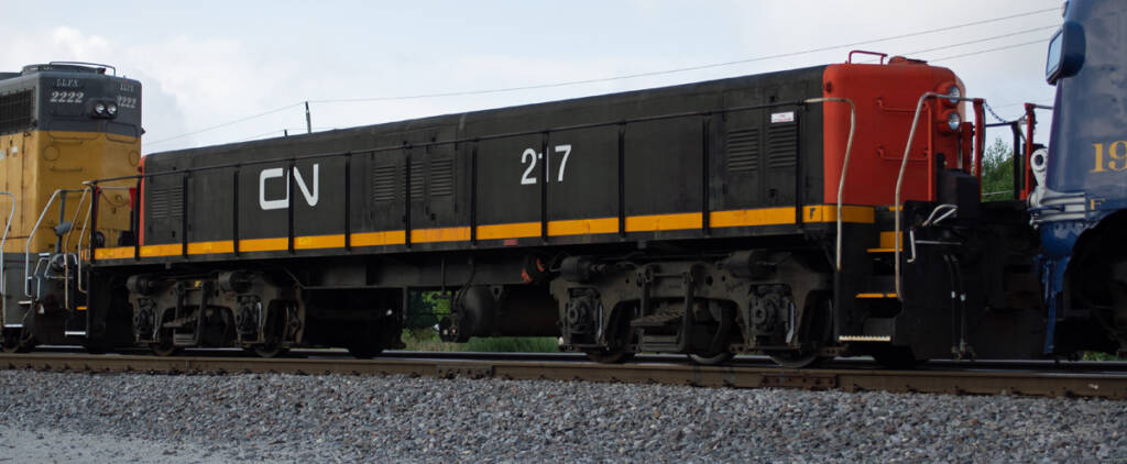 Color photo of four-axle slug unit painted red, yellow, and black with white graphics. A gray-and-yellow EMD hood unit is visible to the left of the slug.