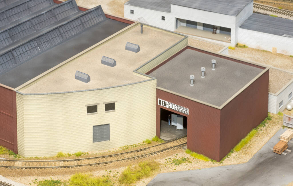 Color photo of tan and brown brick warehouse buildings with gravel roof on scenicked model railroad layout with a cinder block building in the background.