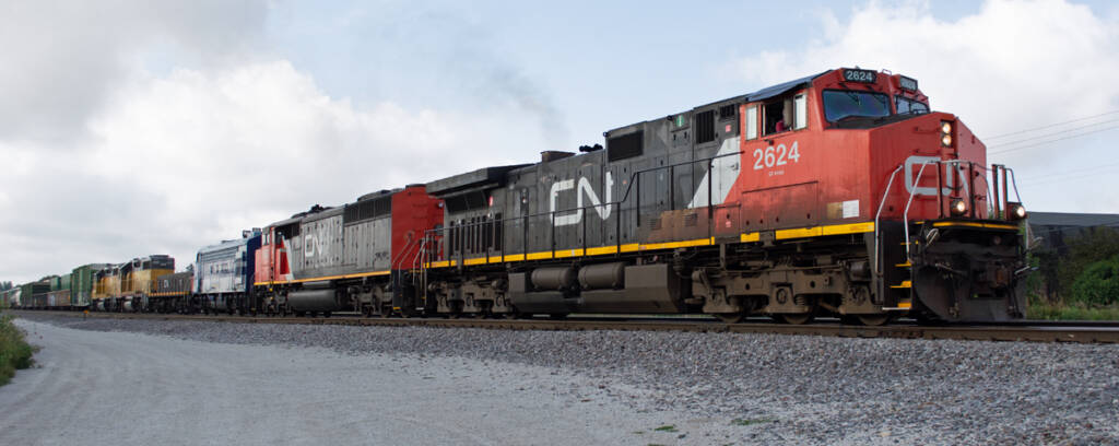 Color photo of train with six diesel locomotives in various paint schemes leading a freight train on a partly cloudy day. A gray gravel road is in the foreground. The track has variegated gray ballast. Green vegetation is visible in the background.