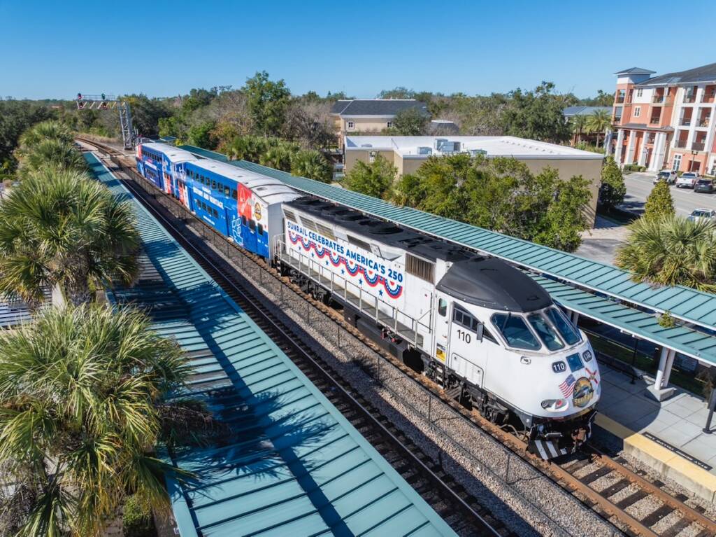White locomotive with red and blue trim and two bilevel passenger cars with red, white, and blue wraps.