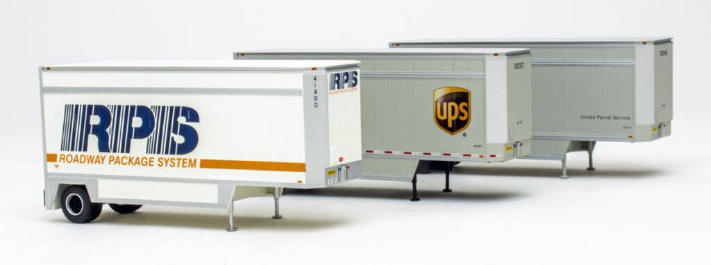 Color photo of three HO scale trailers (one painted white and gray with orange and blue graphics, one painted gray and silver with brown and yellow logo and red-and-white stripes, and one painted gray and silver with black lettering).