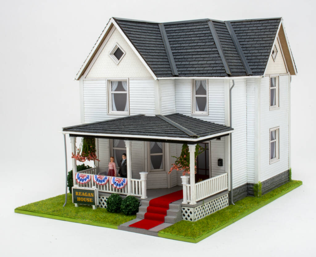 Color photo showing white two-story house with gray shingles and covered front porch with red carpet with and male and female figures. Yard features static grass and small shrubs. Two hanging plants are on the porch.