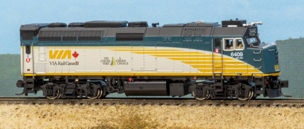Color image of N scale four-axle cowl-body diesel painted green, gray and yellow with red, white, and black graphics on scenicked base with a sky blue background.