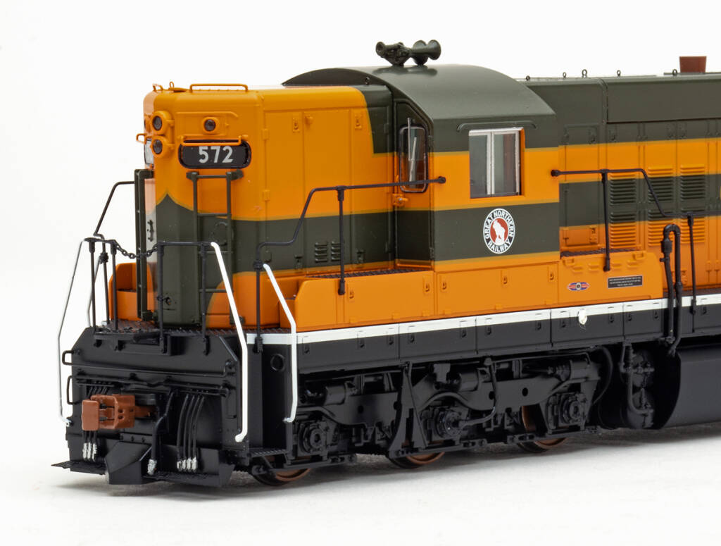 Color photo showing cab and short hood on EMD six-axle hood unit painted orange, green, and yellow with black trucks, sill, and pilot on white background.