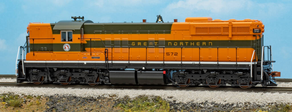 Color photo of six-axle EMD hood unit painted orange and green with yellow stripes and lettering and black trucks and fuel tank on scenicked base with a sky blue background.