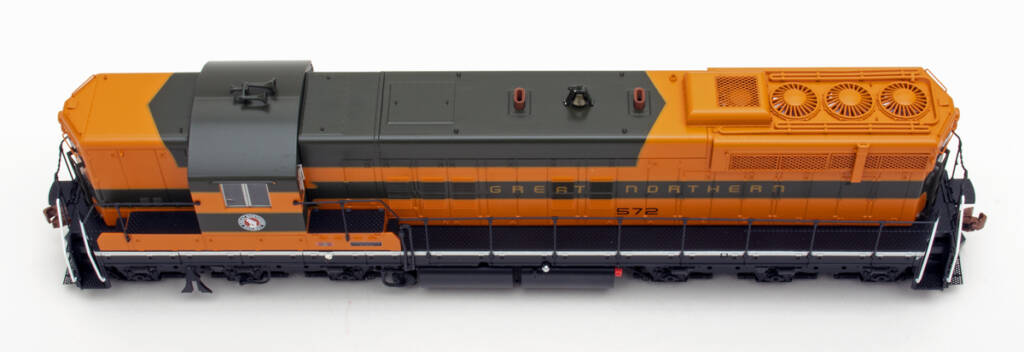 Color photo showing roof of EMD six-axle hood unit painted orange, green, and yellow with black trucks, walkways, sills, and pilots on white background.