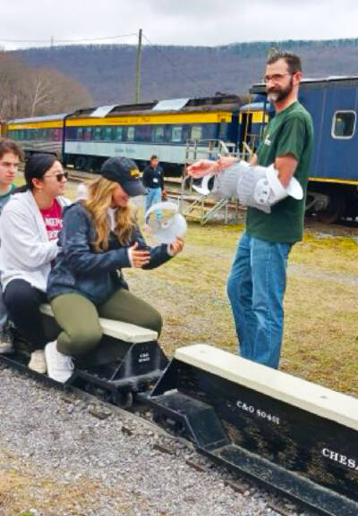 Man handing out paper hats next to small ride-on train. C&O Historical Society George Washington Train Show announced.